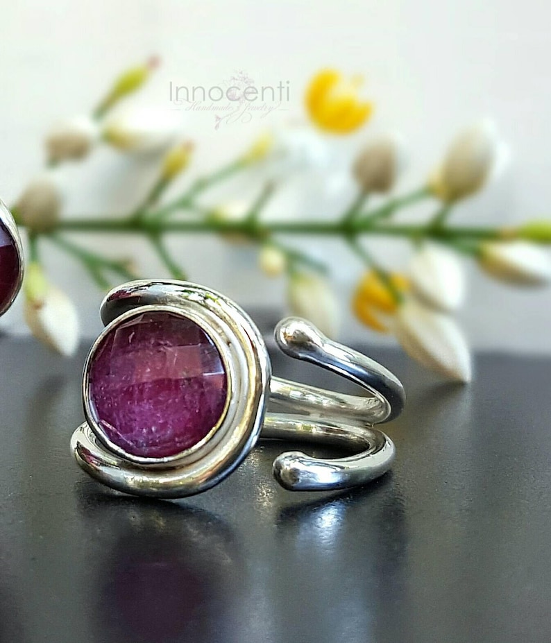 K&ouml;nnte beinhalten: Ein silberner Ring mit einem geschwungenen Design und einem runden, rosafarbenen Edelstein in der Mitte.