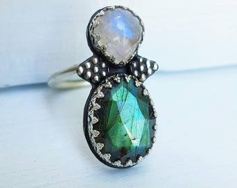 Puede incluir: Un anillo de plata con una piedra preciosa verde iridiscente en forma de pera y una piedra preciosa azul pálido redonda. El anillo tiene un diseño único con detalles decorativos de plata alrededor de las piedras y en la banda.