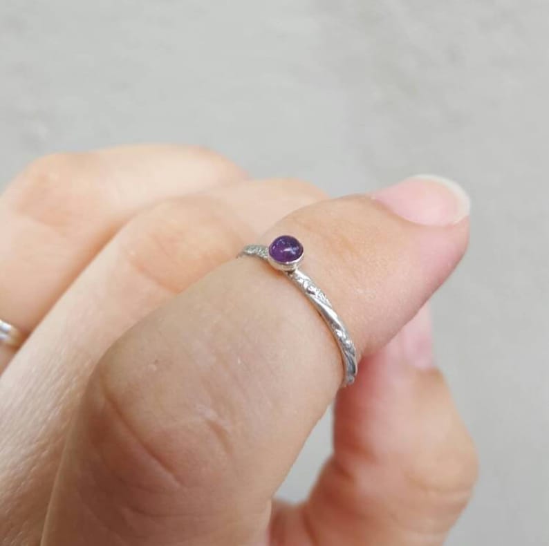 Puede incluir: Un anillo de plata con una peque&ntilde;a gema redonda de color p&uacute;rpura. La banda del anillo tiene un dise&ntilde;o texturizado y grabado. El anillo se muestra en un dedo.