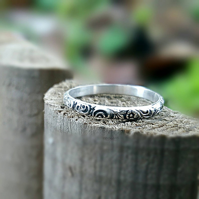 May include: A silver ring with an engraved floral pattern. The ring is set against a blurred background of wood and greenery, highlighting the intricate details of the design. The ring's surface is adorned with delicate flowers and swirling patterns.