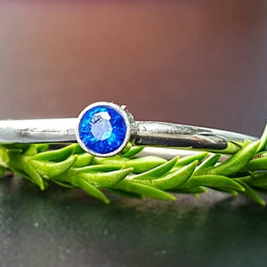 Op de afbeelding: Een zilveren ring met een ronde, helderblauwe edelsteen. De ring ligt op een bedje van groen loof. De achtergrond is een donker, wazig oppervlak. De ring is een sieraad.