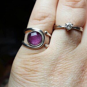 K&ouml;nnte beinhalten: Zwei silberne Ringe an einem Finger. Der Ring unten hat einen violetten Stein, der in eine silberne Fassung gefasst ist. Der Ring oben hat einen einzigen klaren Stein.