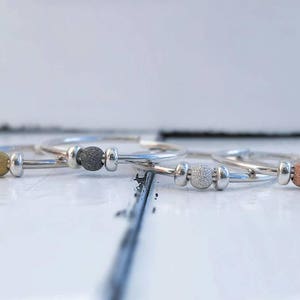 Könnte beinhalten: Vier silberne Ringe, jeder mit einer andersfarbigen Perle: Gold, Dunkelgrau, Silber und Kupfer. Die Perlen sind strukturiert und kugelförmig, was dem schlichten, eleganten Design einen visuellen Reiz verleiht. Die Ringe werden auf einer weißen Oberfläche präsentiert.