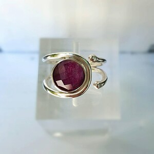 K&ouml;nnte beinhalten: Ein silberner Ring mit einem tiefvioletten Edelstein, der in einer runden Fassung sitzt. Der Ring hat ein einzigartiges, verdrehtes Banddesign.