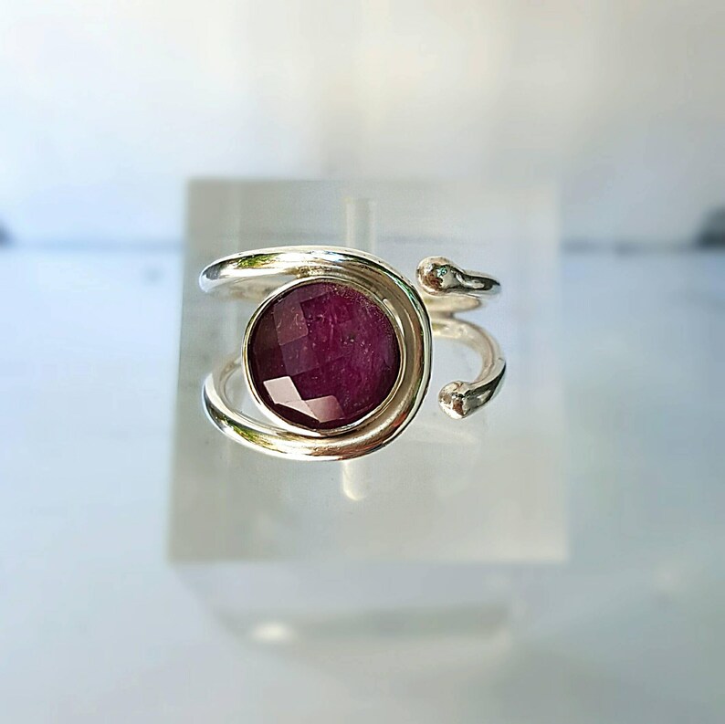 K&ouml;nnte beinhalten: Ein silberner Ring mit einem tiefvioletten Edelstein, der in einer runden Fassung sitzt. Der Ring hat ein einzigartiges, verdrehtes Banddesign.