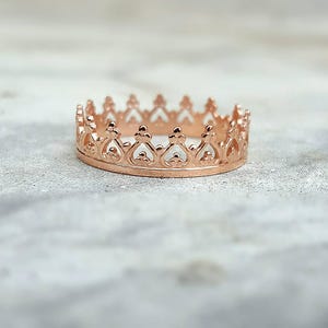 May include: A rose gold crown ring with an intricate design. The ring showcases a series of pointed arches and decorative features, giving it a royal look. The ring is set against a soft, neutral background.