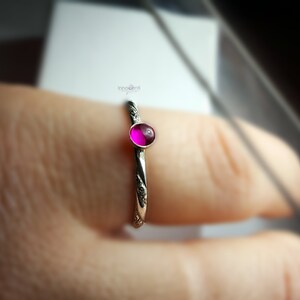 May include: A silver ring featuring a small, round, magenta gemstone. The ring's band is adorned with a subtle floral design. The vibrant gemstone is the central element, set against the detailed silver band. A piece of jewellery.