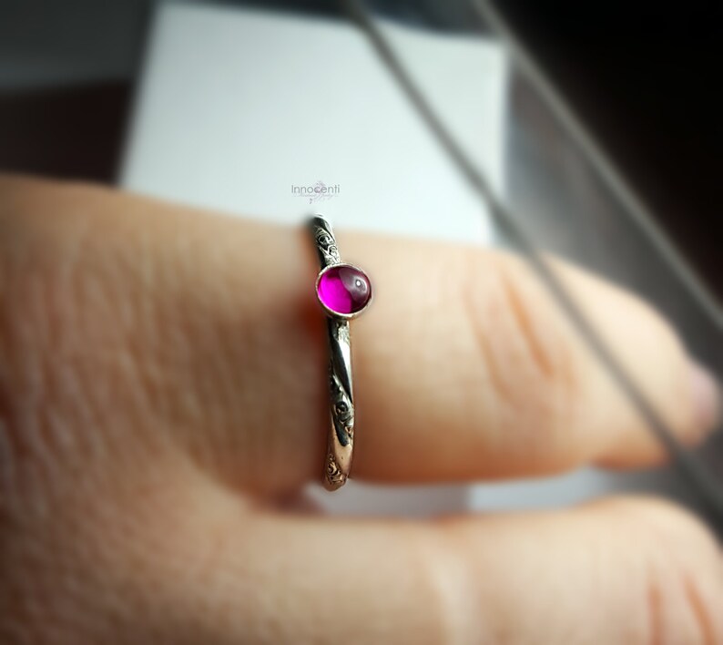 May include: A silver ring featuring a small, round, magenta gemstone. The ring's band is adorned with a subtle floral design. The vibrant gemstone is the central element, set against the detailed silver band. A piece of jewellery.