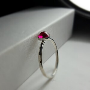 Puede incluir: Un anillo de plata con una banda martillada y una vibrante gema roja y redonda. La gema est&aacute; engastada en un bisel, creando un aspecto elegante y moderno. El anillo se muestra sobre un fondo blanco.