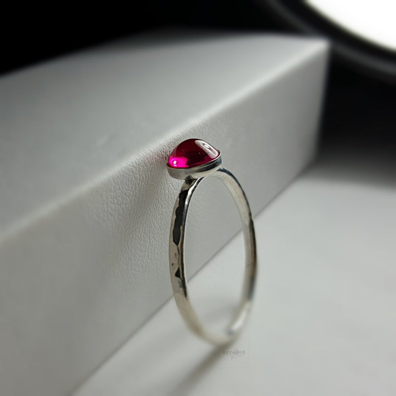 Puede incluir: Un anillo de plata con una banda martillada y una vibrante gema roja y redonda. La gema est&aacute; engastada en un bisel, creando un aspecto elegante y moderno. El anillo se muestra sobre un fondo blanco.