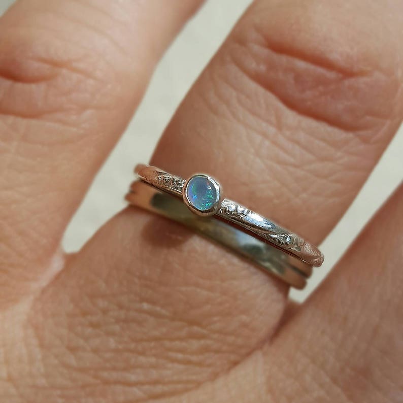 May include: A set of two silver rings. The top ring features a small, round, light blue opal stone. The second ring is a simple band. The top ring has a decorative etched design. The rings are on a finger.