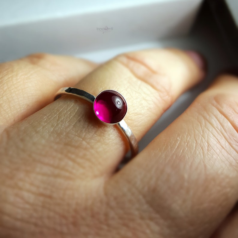 Puede incluir: Un anillo de plata con una banda martillada y una gema redonda de color magenta. La gema es el punto focal, engastada en un dise&ntilde;o simple y elegante. El anillo se muestra en un dedo.