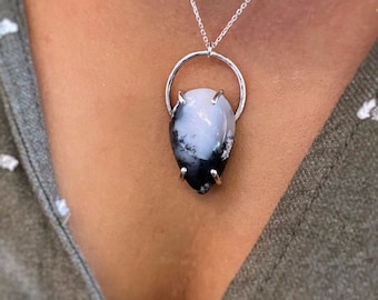 Puede incluir: Un collar con colgante de plata con una piedra en forma de lágrima con patrones en blanco y negro. La piedra está sujeta por un anillo de plata con cuatro garras. El collar se lleva en una cadena de plata.