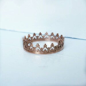 May include: A rose gold crown ring with an intricate, openwork design. The ring features heart-shaped cutouts along the top edge, creating a regal and elegant appearance. The ring is set against a light blue and white background.
