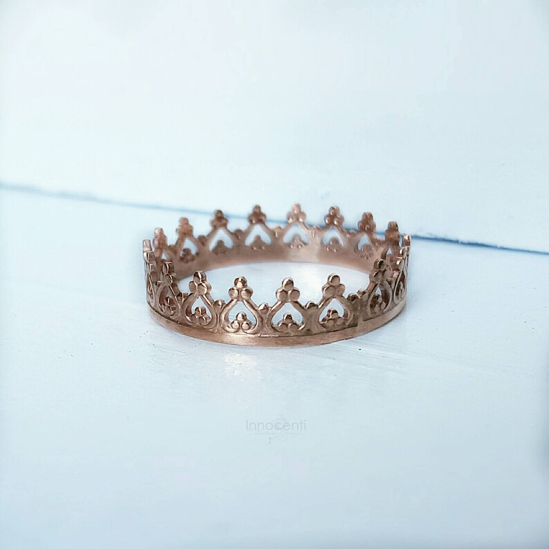 May include: A rose gold crown ring with an intricate, openwork design. The ring features heart-shaped cutouts along the top edge, creating a regal and elegant appearance. The ring is set against a light blue and white background.