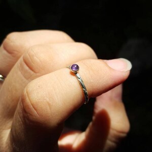 Puede incluir: Un anillo de plata con una peque&ntilde;a gema redonda de color p&uacute;rpura. La banda del anillo tiene un dise&ntilde;o delicado y grabado. El anillo se muestra en un dedo, con un fondo oscuro.