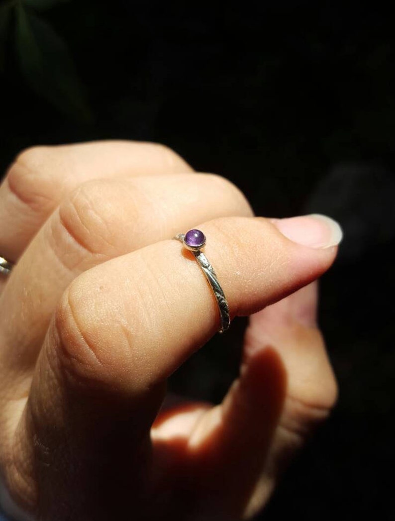Puede incluir: Un anillo de plata con una peque&ntilde;a gema redonda de color p&uacute;rpura. La banda del anillo tiene un dise&ntilde;o delicado y grabado. El anillo se muestra en un dedo, con un fondo oscuro.