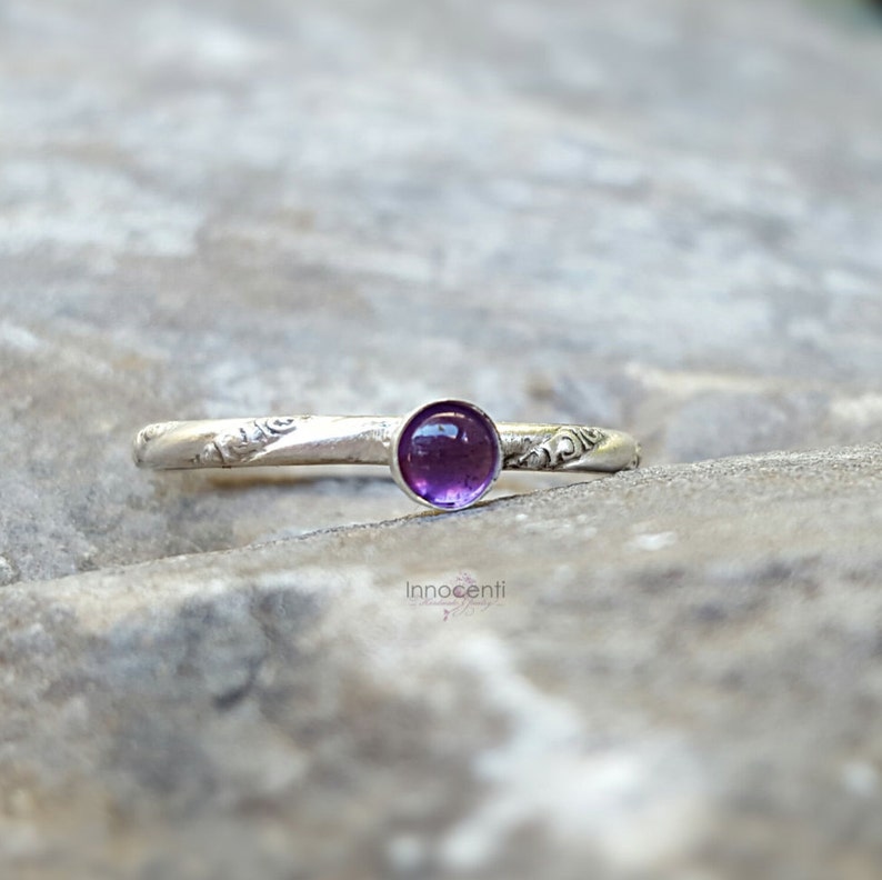 Puede incluir: Un anillo de plata con una piedra preciosa redonda de color p&uacute;rpura intenso. La banda presenta un dise&ntilde;o floral grabado. El anillo se encuentra sobre un fondo neutro y texturizado, resaltando los detalles de la joya.