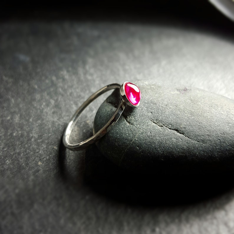 Puede incluir: Un anillo de plata con una banda martillada y una vibrante gema roja de forma ovalada. El anillo se muestra sobre un fondo oscuro y texturizado, resaltando el color de la gema y el dise&ntilde;o del anillo. El anillo es una pieza de joyer&iacute;a.