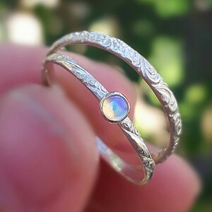 May include: A silver ring with an opal gemstone. The ring features a double band design with intricate floral detailing. The opal is round and displays a mix of iridescent colors. The ring is held up against a blurred green background.