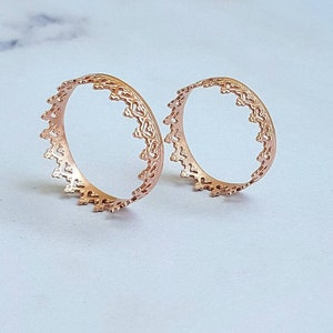 May include: Two rose gold open-work rings with a delicate, ornate design. The rings have a circular shape with a detailed, repeating pattern along the outer edge. The rings are displayed on a light-coloured surface.