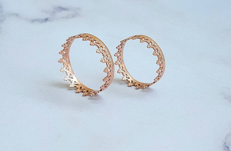 May include: Two rose gold open-work rings with a delicate, ornate design. The rings have a circular shape with a detailed, repeating pattern along the outer edge. The rings are displayed on a light-coloured surface.