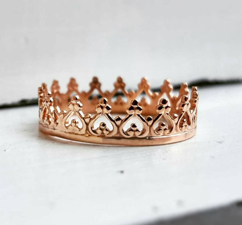 May include: A rose gold crown ring with detailed filigree. The ring has heart-shaped cutouts and decorative, pointed arches. The ring is set against a white background, emphasising its delicate design. The ring's diameter is approximately 2 cm.