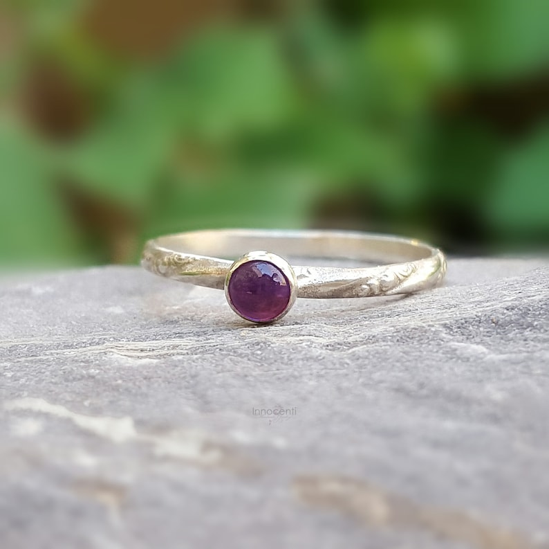 Puede incluir: Un anillo de plata con una peque&ntilde;a gema redonda de color p&uacute;rpura oscuro. La banda tiene un delicado patr&oacute;n floral grabado. El anillo se encuentra sobre un fondo verde y gris borroso, resaltando la simple elegancia de la joya.