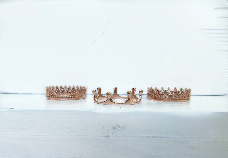 May include: Three rose gold crown rings are displayed on a white surface. The crowns have different designs, with one in the centre featuring a unique, curved shape. The word "Innocenti" is visible in the image.