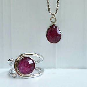 K&ouml;nnte beinhalten: Ein Schmuckset mit einem silbernen Ring und einem Anh&auml;nger mit einem tief roten Edelstein. Der Ring hat ein verdrehtes Band und der Anh&auml;nger ist tropfenf&ouml;rmig.