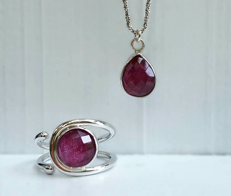K&ouml;nnte beinhalten: Ein Schmuckset mit einem silbernen Ring und einem Anh&auml;nger mit einem tief roten Edelstein. Der Ring hat ein verdrehtes Band und der Anh&auml;nger ist tropfenf&ouml;rmig.