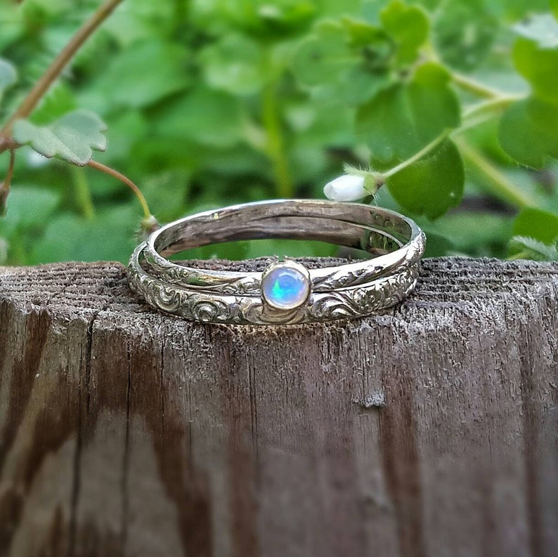 May include: Two silver rings with intricate floral patterns. One ring features a small, round, opalescent gemstone. The rings are stacked on a weathered wooden surface, with a blurred green background of foliage.