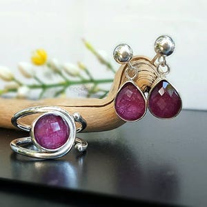K&ouml;nnte beinhalten: Ein Set aus silbernen Ohrringen und einem Ring mit rosafarbenen Edelstein-Akzenten. Die Ohrringe sind tropfenf&ouml;rmig und der Ring ist ein schlichtes Band mit einem runden Stein.