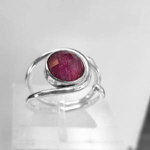 K&ouml;nnte beinhalten: Ein silberner Ring mit einem tief roten Edelstein, der in einem verdrehten Band gefasst ist.