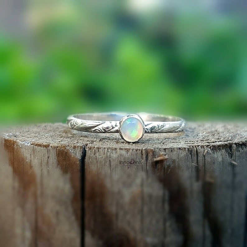 May include: A silver ring with an oval opal gemstone. The ring band has a delicate floral pattern. The opal has a milky white base with flashes of color. The ring is displayed on a wooden surface.