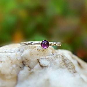 Puede incluir: Un anillo de plata con una peque&ntilde;a gema redonda y p&uacute;rpura. La banda presenta un dise&ntilde;o floral grabado. El anillo se encuentra sobre un fondo verde y blanco borroso, resaltando los delicados detalles de la joya.