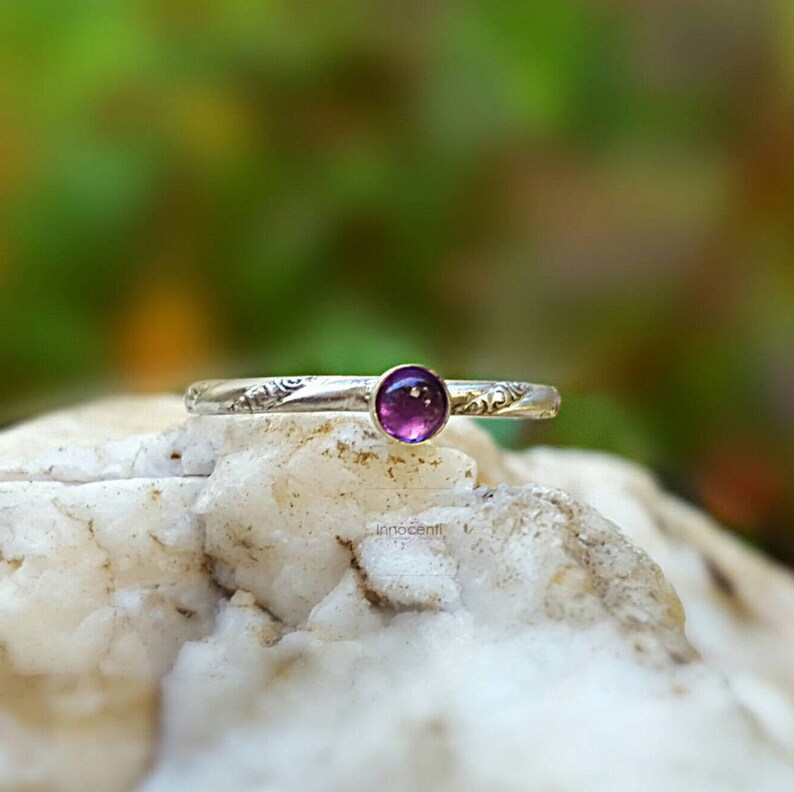 Puede incluir: Un anillo de plata con una peque&ntilde;a gema redonda y p&uacute;rpura. La banda presenta un dise&ntilde;o floral grabado. El anillo se encuentra sobre un fondo verde y blanco borroso, resaltando los delicados detalles de la joya.