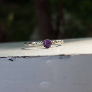 Puede incluir: Un anillo de plata con una gema redonda de color p&uacute;rpura intenso. La banda tiene un dise&ntilde;o floral texturizado. El anillo est&aacute; sobre una superficie blanca con un fondo verde borroso.