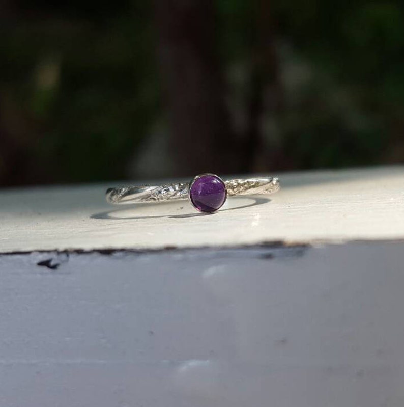 Puede incluir: Un anillo de plata con una gema redonda de color p&uacute;rpura intenso. La banda tiene un dise&ntilde;o floral texturizado. El anillo est&aacute; sobre una superficie blanca con un fondo verde borroso.