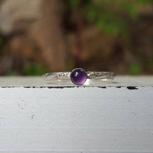 Puede incluir: Un anillo de plata con una banda grabada y una piedra preciosa redonda de color p&uacute;rpura intenso. El anillo se encuentra sobre una superficie blanca con un fondo borroso verde y marr&oacute;n, mostrando el delicado dise&ntilde;o y contraste de color.