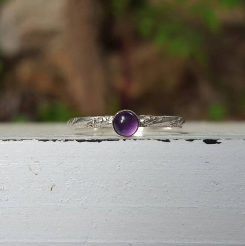 Puede incluir: Un anillo de plata con una banda grabada y una piedra preciosa redonda de color p&uacute;rpura intenso. El anillo se encuentra sobre una superficie blanca con un fondo borroso verde y marr&oacute;n, mostrando el delicado dise&ntilde;o y contraste de color.