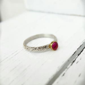 Può includere: Un anello d'argento con castone dorato che tiene una gemma rossa rotonda. La fascia presenta un motivo floreale inciso. L'anello è appoggiato su una superficie di legno bianco.