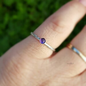 Puede incluir: Un anillo de plata con una peque&ntilde;a gema redonda de color p&uacute;rpura. La banda tiene un dise&ntilde;o delicado y grabado. El anillo se muestra en un dedo, con un fondo borroso.