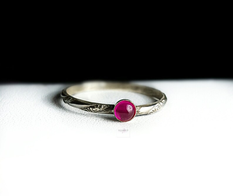May include: A silver ring with an engraved floral design. It has a round, magenta gemstone set in the centre. The ring is displayed on a white surface with a black backdrop.