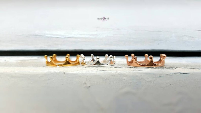 May include: Three small crown-shaped rings in gold, silver, and rose gold. The rings are displayed on a white surface. The crowns have five points each and are likely jewelry.