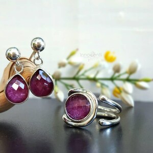 K&ouml;nnte beinhalten: Ein Set aus silbernen Ohrringen und einem Ring mit rosafarbenen Edelstein-Akzenten. Die Ohrringe sind tropfenf&ouml;rmig und der Ring hat einen runden Edelstein.