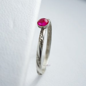 Op de afbeelding: Een zilveren ring met een kleine, ovale, helder roze edelsteen. De band heeft een textuur, geëtst ontwerp. De ring staat tegen een witte achtergrond, waardoor de details van het sieraad worden benadrukt. De ring is een delicaat sieraad.