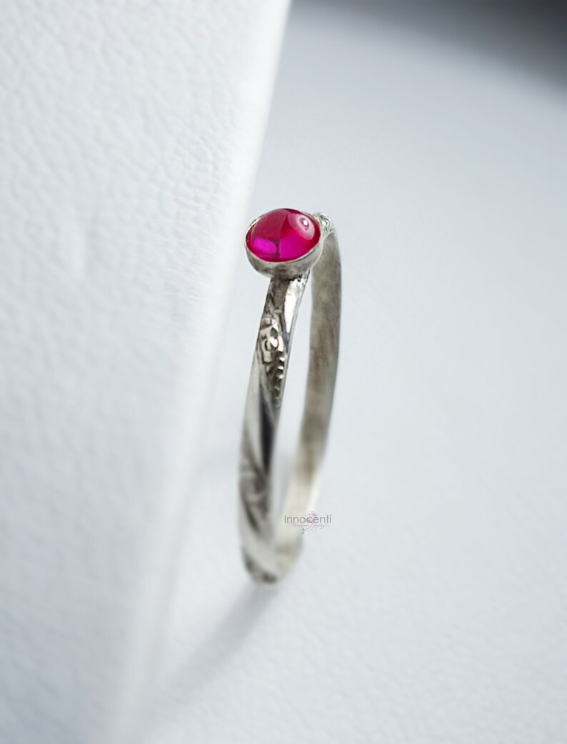 May include: A silver ring featuring a small, oval, bright pink gemstone. The band has a textured, etched design. The ring is set against a white backdrop, highlighting the jewellery's details. The ring is a delicate piece of jewellery.
