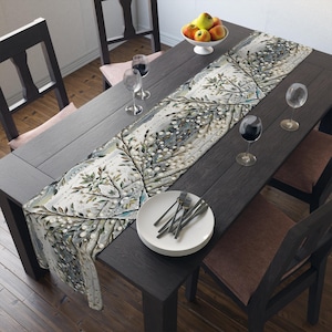 May include: A white table runner with a floral design featuring white flowers and green leaves on a gray background. The runner is placed on a dark wood table with white plates and silverware.