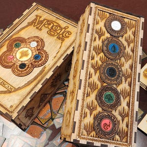 May include: Wooden deck boxes for Magic: The Gathering cards. The boxes are light brown with intricate laser-cut designs and the words "Magic" and "Deck Box". The boxes have circular tokens with various symbols and colors. Several game cards are scattered around the boxes.